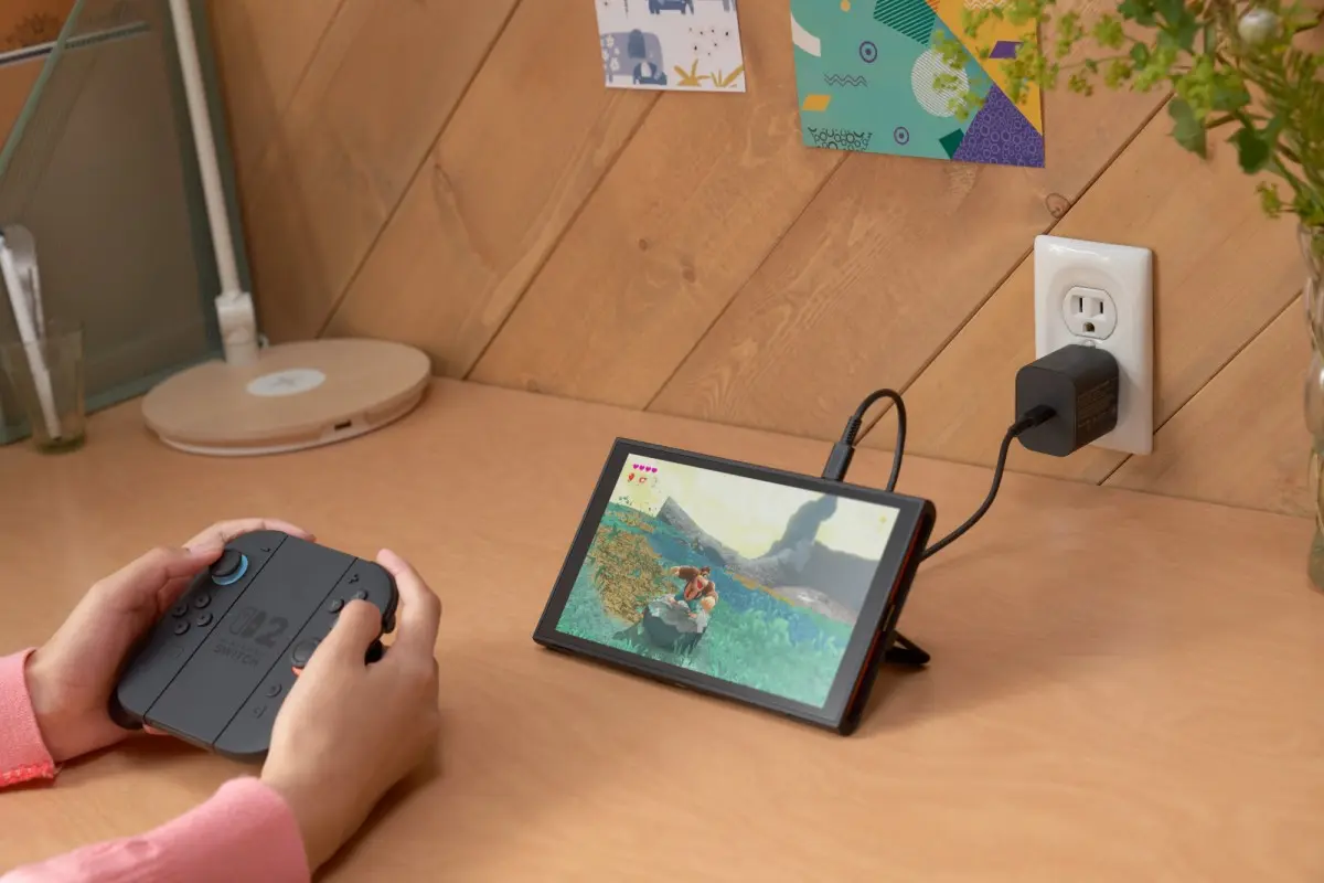 دست‌های بازیکن در حال استفاده از Joy-Con های جداشده Nintendo Switch 2 و بازی در حال اجرا روی صفحه‌نمایش کنسول که روی میز قرار گرفته و به شارژر متصل است