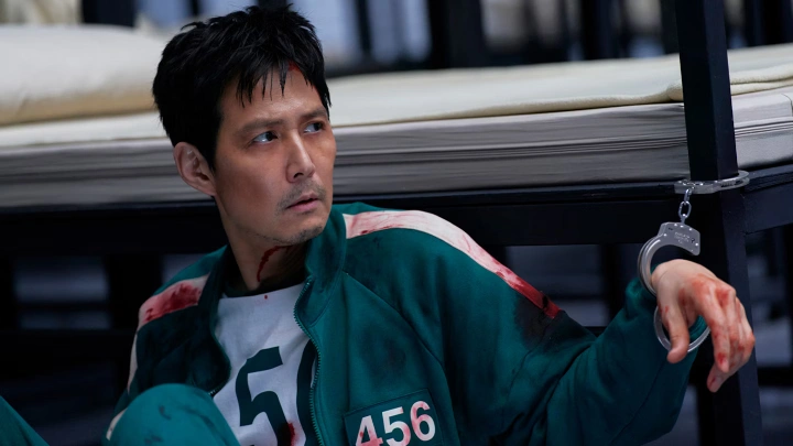 لی جونگ‌جه در نقش گی‌هون در فصل سوم سریال Squid Game، با لباس خون‌آلود و دست‌بندزده در صحنه‌ای پرتنش