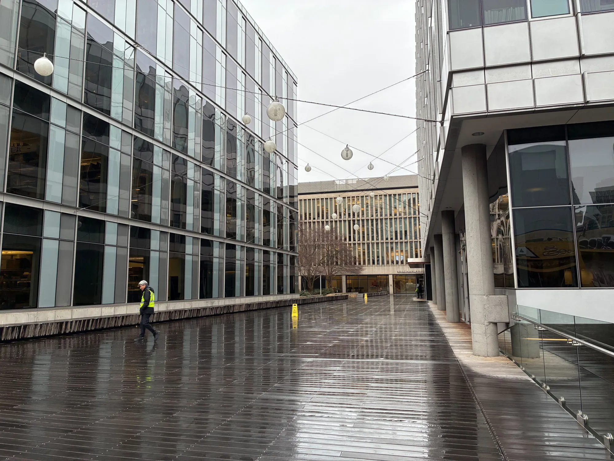 نمونه عکس روز با آیفون 16e از خیابانی بارانی با ساختمان‌های شیشه‌ای و بازتاب نور طبیعی روی زمین مرطوب